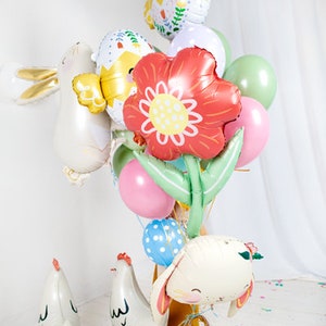 May include: A collection of colorful Easter-themed balloons, including a chick, a bunny, a flower, and an egg. The balloons are arranged in a bouquet and are tied together with a ribbon. There are also two small white chicks on the ground.
