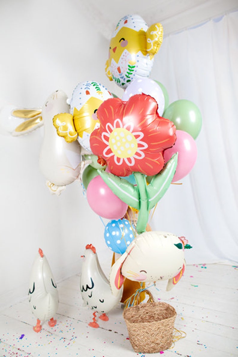 May include: A collection of colorful Easter-themed balloons, including a chick, a bunny, a flower, and an egg. The balloons are arranged in a bouquet and are tied together with a ribbon. There are also two small white chicks on the ground.