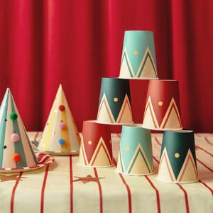 Op de afbeelding: Een stapel kleurrijke papieren bekers met geometrische ontwerpen in teal, rood en donkergroen, met een gouden stip accent. Feestmutsen met pompons en sterrenaccenten zijn ook zichtbaar. De bekers en hoeden staan op een gestreepte tafelkleed.