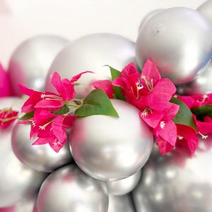 May include: A cluster of silver balloons adorned with vibrant pink bougainvillea flowers and green leaves. The balloons are arranged in a layered composition, with a few pink and blue balloons visible in the background.