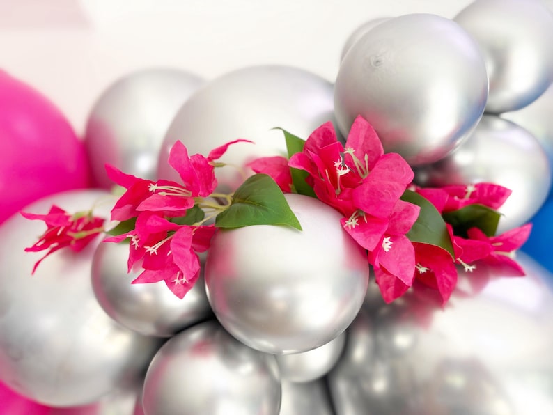 May include: A cluster of silver balloons adorned with vibrant pink bougainvillea flowers and green leaves. The balloons are arranged in a layered composition, with a few pink and blue balloons visible in the background.