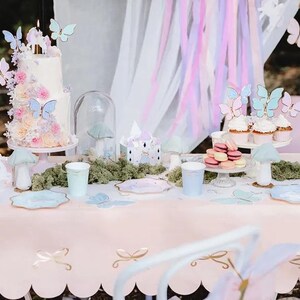 May include: A pink table with a scalloped edge is set for a party with pastel blue and pink butterfly themed paper plates, cups, and napkins. A two-tiered cake decorated with butterflies and flowers sits on a white cake stand. There are also cupcakes with butterfly toppers and macarons on a white cake stand.