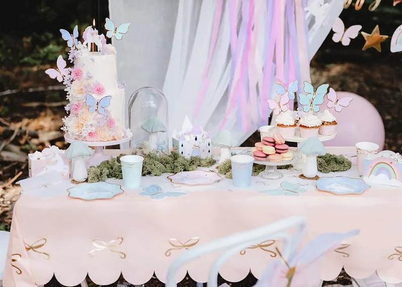 May include: A pink table with a scalloped edge is set for a party with pastel blue and pink butterfly themed paper plates, cups, and napkins. A two-tiered cake decorated with butterflies and flowers sits on a white cake stand. There are also cupcakes with butterfly toppers and macarons on a white cake stand.