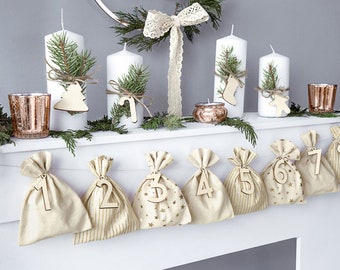 May include: A festive Christmas display featuring white pillar candles adorned with greenery and wooden ornaments. A garland of greenery and small copper-colored candle holders are also present. Below, numbered fabric bags create an advent calendar.