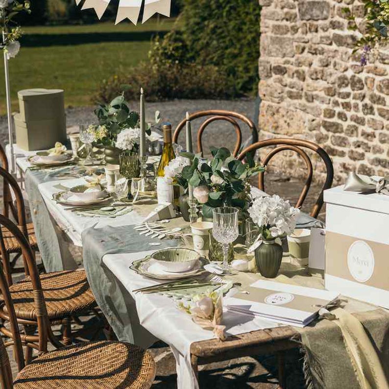 May include: A beautifully set outdoor dining table with a white tablecloth, adorned with floral arrangements, candles, and elegant tableware. The table setting includes plates, glasses, and decorative elements, creating a sophisticated ambiance. Wicker chairs surround the table.