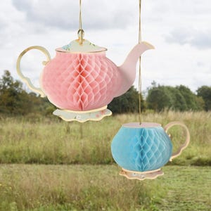 May include: Two hanging paper honeycomb decorations shaped like teapots. One is pink with a light pink spout and handle, the other is blue with a light pink handle. Both have floral trim and are suspended by gold strings.