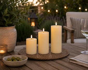 May include: Three ivory-colored pillar candles of varying heights, each with a lit flame, sit on a round woven placemat. The candles are on a wooden table, with a bowl of olives and a wine glass visible. The scene is set outdoors.