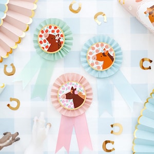 Pode incluir: Três rosetas de papel azul e rosa com um desenho de cavalo e ferraduras douradas espalhadas sobre um fundo xadrez azul e branco.