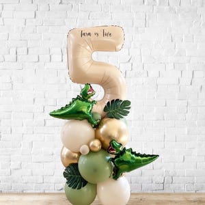 May include: A balloon arrangement for a fifth birthday. The arrangement includes a large beige number 5 balloon with the text "Tara is Five". Green dinosaur balloons and other balloons in shades of green, gold, and beige are also included.