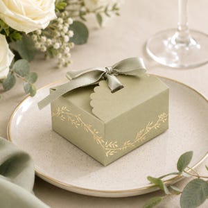 May include: A sage green gift box with a scalloped edge and a matching ribbon bow. The box is decorated with gold leaf branch designs and sits on a speckled plate. White roses and greenery are in the background.