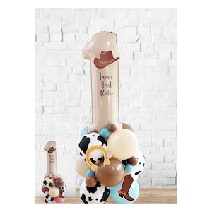 Könnte beinhalten: Ein großer brauner Ballon in Form der Zahl Eins mit einem Cowboyhut oben drauf. Der Ballon trägt den Text "Tara's First Rodeo". Der Ballon ist von kleineren Ballons in verschiedenen Farben und Mustern umgeben, darunter Kuhmuster, Braun, Blau und Beige.