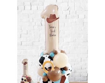 May include: A large brown number one balloon with a cowboy hat on top. The balloon has the text "Tara's First Rodeo" printed on it. The balloon is surrounded by smaller balloons in various colors and patterns, including cow print, brown, blue, and beige.