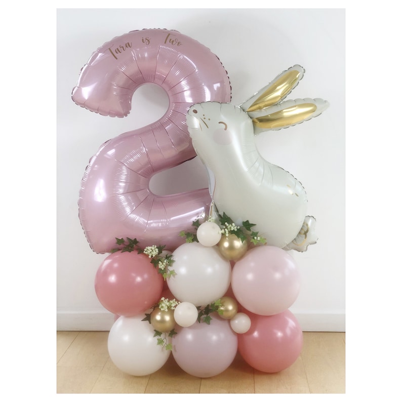 May include: A pink number two balloon with the text "Tara is two" on it. A white bunny balloon is next to the number two balloon. The balloons are surrounded by smaller pink, white, and gold balloons.