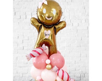 Może przedstawiać: Złoty balon w kształcie piernikowego ludzika z różową kokardą, sercem z imieniem "Tara" i podstawą z różowych, białych i złotych balonów. Dołączone są dwa balony w kształcie cukierków.