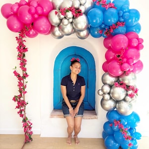 May include: A vibrant balloon arch in shades of pink, silver, and blue, adorned with pink flowers. The arch frames a blue arched doorway against a white wall. A person is sitting in the doorway.
