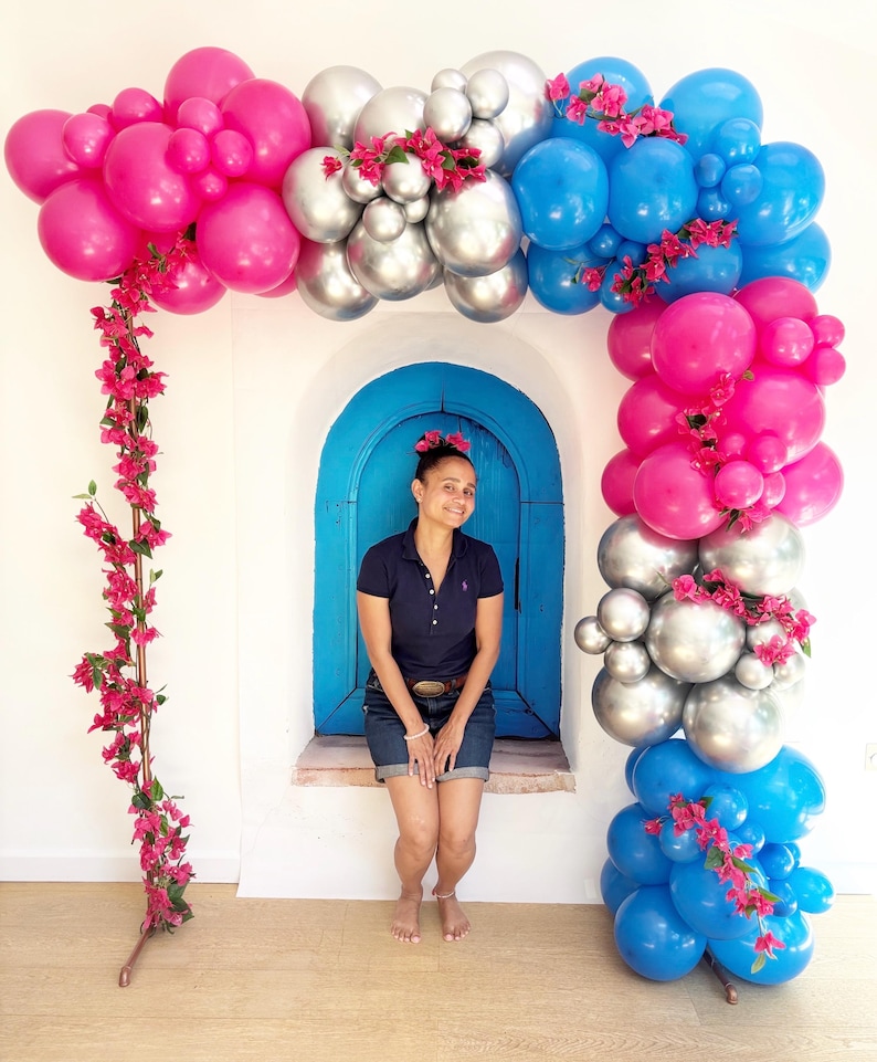 May include: A vibrant balloon arch in shades of pink, silver, and blue, adorned with pink flowers. The arch frames a blue arched doorway against a white wall. A person is sitting in the doorway.