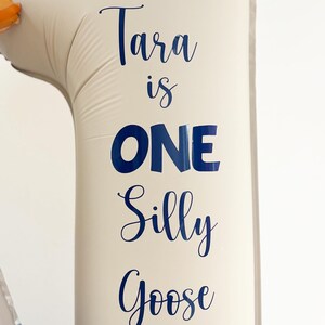 May include: A beige number one balloon with the words "Tara is ONE Silly Goose" in dark blue script. The balloon is inflated and suitable for a birthday party. The background is plain white.