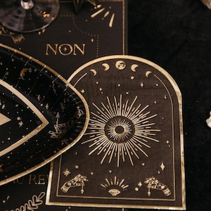 May include: Black and gold paper party plate with a celestial design. The plate features a sun with a radiating starburst, moons, stars, and a hand with an eye. The text "NON" is visible in the background.