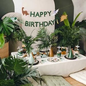 May include: A jungle-themed birthday party table setting with green plants, a white tablecloth, and a banner that reads "Happy Birthday". The table is decorated with dinosaur-themed plates, cups, and party hats.