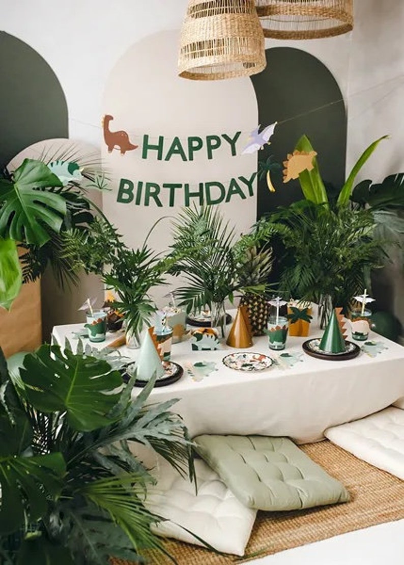 May include: A jungle-themed birthday party table setting with green plants, a white tablecloth, and a banner that reads "Happy Birthday". The table is decorated with dinosaur-themed plates, cups, and party hats.