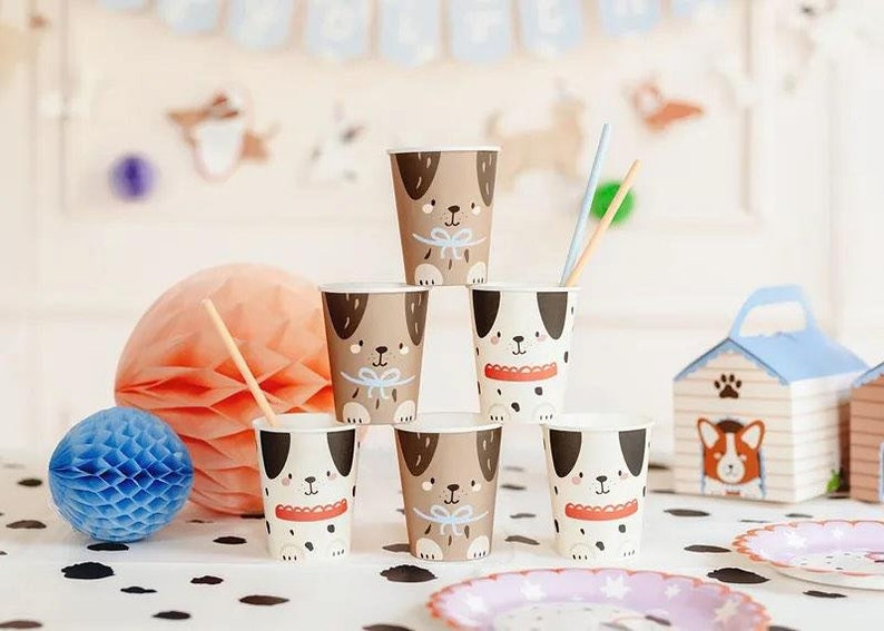Puede incluir: Conjunto de seis vasos de papel con diferentes ilustraciones de perros. Los vasos son marrones y blancos con detalles negros. Los perros llevan diferentes accesorios, como una pajarita, un collar con una etiqueta y una bandana roja.