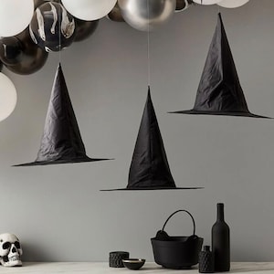 May include: Three black witch hats suspended from above, with a wide brim and pointed crown. The hats are part of a Halloween-themed display, with black and white balloons in the background. Other Halloween decorations include a skull, cauldron, and bottles.