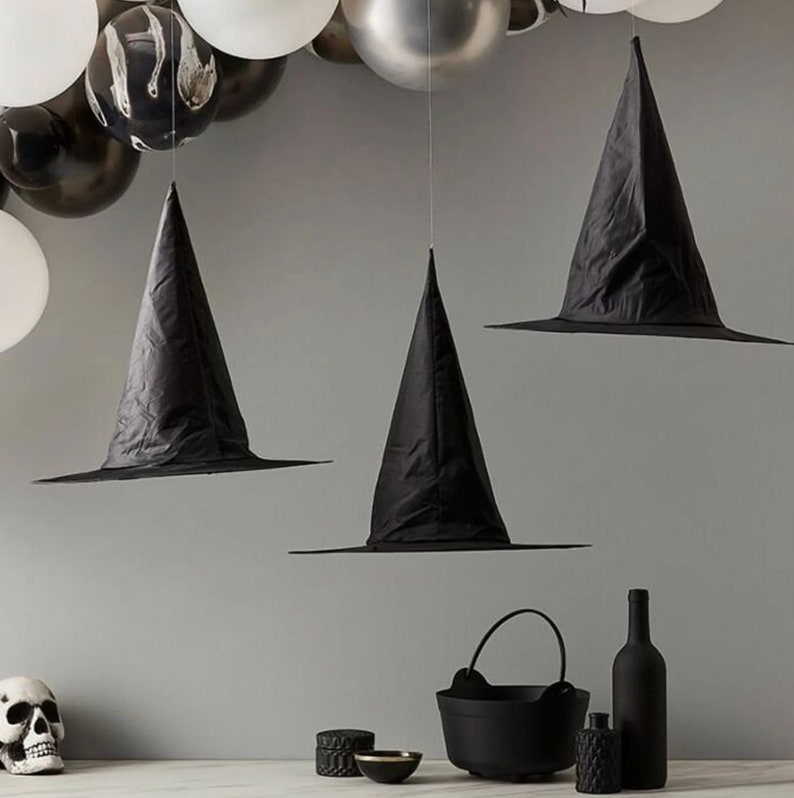 May include: Three black witch hats suspended from above, with a wide brim and pointed crown. The hats are part of a Halloween-themed display, with black and white balloons in the background. Other Halloween decorations include a skull, cauldron, and bottles.