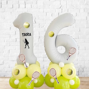Puede incluir: Un arreglo de globos festivo que deletrea el número 16, perfecto para un cumpleaños con temática de tenis. Los globos son de color gris claro, con pegatinas de pelotas y raquetas de tenis. El número 1 tiene el nombre "TARA" y una silueta de tenista.