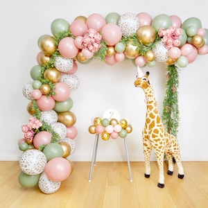 May include: A green, pink and gold balloon arch with pink flowers and greenery. A stuffed giraffe wearing a party hat stands in front of the arch.