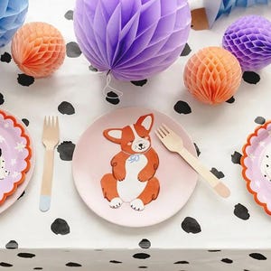 Puede incluir: Una mesa festiva con platos con ilustraciones de perros, tenedores de madera y decoraciones de panal de papel en tonos azules, naranjas y morados. El mantel es blanco con lunares negros, creando un ambiente l&uacute;dico.