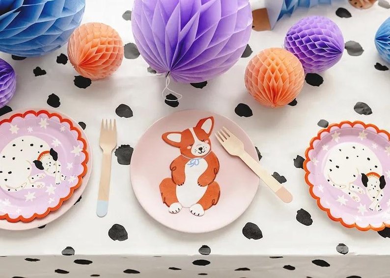 Puede incluir: Una mesa festiva con platos con ilustraciones de perros, tenedores de madera y decoraciones de panal de papel en tonos azules, naranjas y morados. El mantel es blanco con lunares negros, creando un ambiente l&uacute;dico.