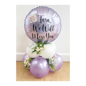 Könnte beinhalten: Ballonarrangement mit einem großen, runden, lila Ballon mit der Aufschrift "Tara, We Will Miss You". Das Arrangement umfasst weiße und lila Ballons, Grünzeug und dekorative Schmetterlinge. Eine weiße Rose schmückt den oberen Ballon.