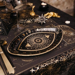 May include: Black and gold paper plate with an eye design. The plate is decorated with stars, moons, and other celestial elements. The plate is part of a set of Halloween party supplies.