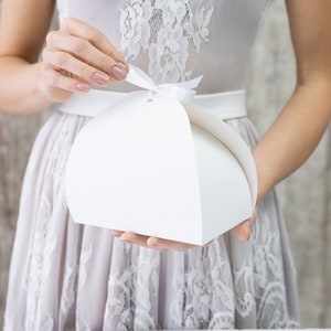 Puede incluir: Una caja de regalo blanca semicircular con una cinta a juego es sostenida por una persona. La caja está hecha de papel grueso y tiene un diseño limpio y minimalista. La persona lleva un vestido de color claro.
