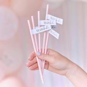 May include: A bundle of pink paper straws with white labels that read "TEAM bride" in a script font. The straws are held in a hand with a light pink manicure. The background is a soft, blurred pink.