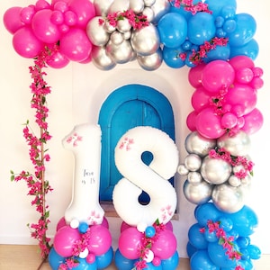 May include: A vibrant balloon arch in shades of pink, silver, and blue, adorned with pink flowers. White number balloons, one and eight, are at the base, with the text "Tara is 18" on the number one balloon. The arch frames a blue door.