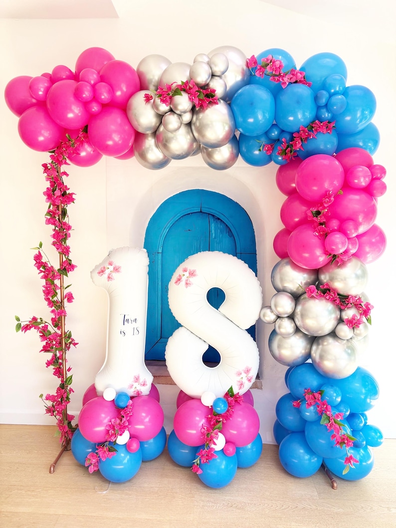 May include: A vibrant balloon arch in shades of pink, silver, and blue, adorned with pink flowers. White number balloons, one and eight, are at the base, with the text "Tara is 18" on the number one balloon. The arch frames a blue door.