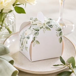 May include: A white, square gift box decorated with a floral pattern of white flowers and green leaves. The box is on a white plate with a gold rim, beside a green napkin and a vase of white flowers.