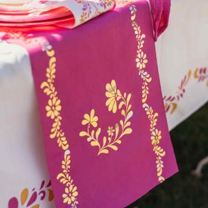 May include: A magenta table runner with gold floral designs. The runner features a central floral motif and borders of stylized flowers and leaves. The runner is placed on a white tablecloth with a matching floral pattern.