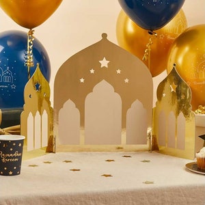 K&ouml;nnte beinhalten: Goldfarbene Papierausstanzungen in Form von Moscheen mit Sternaussparungen, angeordnet auf einer wei&szlig;en Tischdecke. Goldene und blaue Luftballons im Hintergrund. Ein Pappbecher mit der Aufschrift "Ramadan Kareem" ist zu sehen.