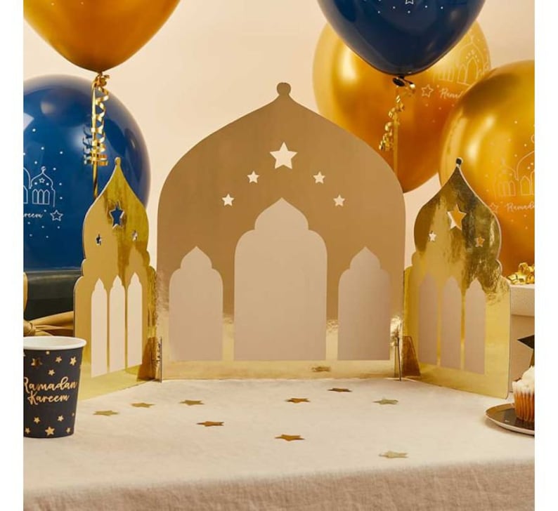K&ouml;nnte beinhalten: Goldfarbene Papierausstanzungen in Form von Moscheen mit Sternaussparungen, angeordnet auf einer wei&szlig;en Tischdecke. Goldene und blaue Luftballons im Hintergrund. Ein Pappbecher mit der Aufschrift "Ramadan Kareem" ist zu sehen.