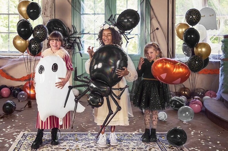 May include: Three children dressed in Halloween costumes hold large inflatable decorations. A girl in a red and white striped dress holds a white ghost balloon. A girl in a white dress holds a black spider balloon. A girl in a black dress holds a pumpkin balloon.