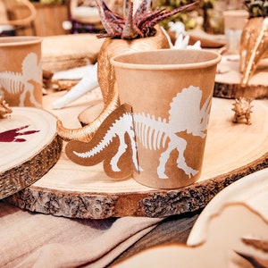 May include: Brown paper cups featuring a white dinosaur skeleton design. The cups are placed on a wooden surface, with a decorative dinosaur tail attached to one. Other wooden decorations and a beige tablecloth are also visible.