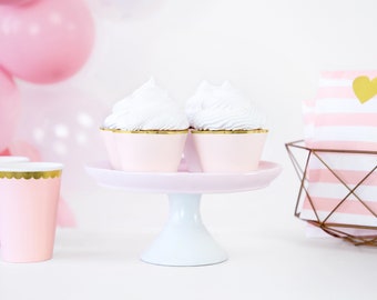 May include: Two white cupcakes with pink frosting on a white cake stand with a pink base. The cupcakes are on a white surface with pink and white striped napkins in a gold wire basket.