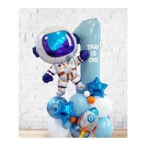 May include: A festive balloon arrangement featuring an astronaut, a number one, and star-shaped balloons. The astronaut balloon is white and orange with a blue visor. The number one balloon is light blue, with the text "TARA IS ONE".