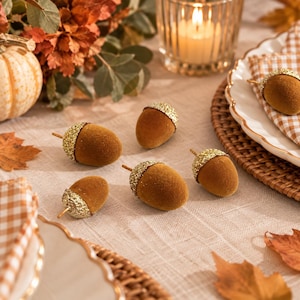 May include: A collection of five decorative acorns featuring brown velvet-textured bodies and gold glitter-covered caps. Each acorn has a small stem protruding from the top, arranged on a light-coloured fabric surface with a fall-themed table setting.