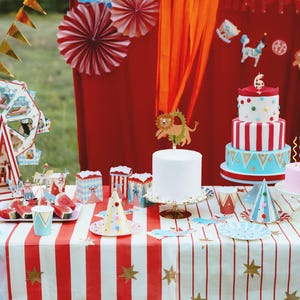 Op de afbeelding: Een feesttafel met circusthema met een rood-wit gestreept tafelkleed. Decoraties zijn onder meer een reuzenrad, feesthoeden, taarten en een banner. De taarten zijn versierd met rood-witte strepen en kleurrijke accenten.