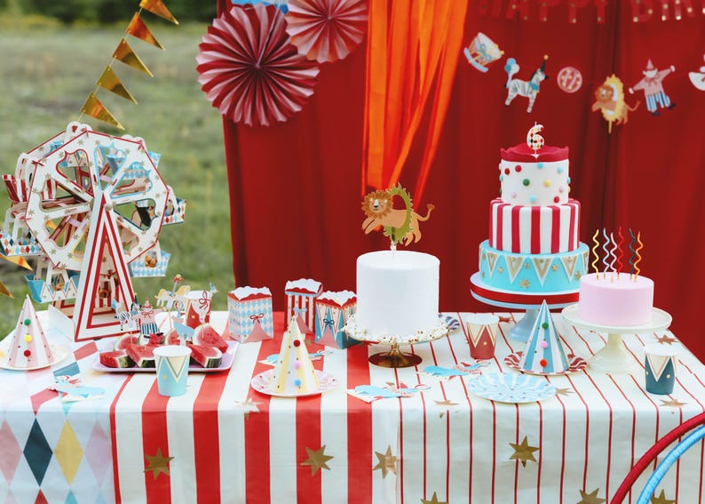 Op de afbeelding: Een feesttafel met circusthema met een rood-wit gestreept tafelkleed. Decoraties zijn onder meer een reuzenrad, feesthoeden, taarten en een banner. De taarten zijn versierd met rood-witte strepen en kleurrijke accenten.