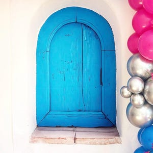 Peut inclure: Une porte en bois bleu vif encastrée dans un mur blanc, encadrée par une ouverture arquée. La porte présente un grain de bois visible et un aspect vieilli. Des ballons roses, argentés et bleus décorent le haut et le côté du mur, suggérant une fête.