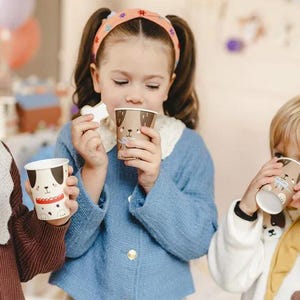 Puede incluir: Tres ni&ntilde;os beben de vasos de papel coloridos con ilustraciones de perros. Los vasos est&aacute;n decorados con la cara de un perro marr&oacute;n y blanco y una nariz roja. Los ni&ntilde;os llevan ropa informal y sonr&iacute;en.